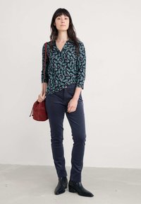 Bluza tamno cvjetnog uzorka s 3/4 rukavima, u kombinaciji s tamnoplavim uskim hlačama i crnim čizmama na petu do gležnja. Burandi torba u obliku kante.