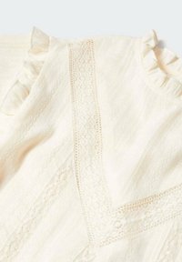 Hellbeige Bluse mit Rüschendetails, strukturiertem Stoff und Spitzenverzierungen entlang der Mitte. Weicher, zarter Look mit dezenten Mustern.