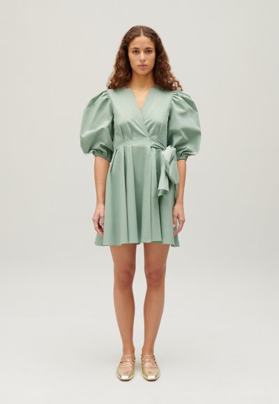 Rochie înfofoficată din material verde deschis, cu mâneci bufante, talie ajustată și detaliu cu fundiță; fustă până la genunchi. Modelul poartă pantofi aurii.