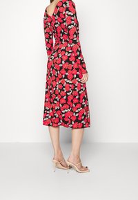 Kvinna klädd i en svart midiklänning med röd blommig tryck, långärmad, och beige högklackade sandaler, stående i profil mot en vit bakgrund.