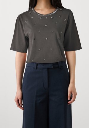 Žena nosící tmavě šedé tričko s krátkými rukávy ozdobené malými květinovými korálkovými detaily, doplněné o tmavě modré kalhoty.