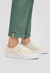 Hellbeige Leinensneaker mit weißen Gummisohlen. Merkmale sind die abgerundete Form, ein Schnürdesign und ein dezentes Markenschild an der Seite. Grüne, hochgekrempelte Hose.