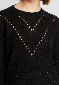 Nahaufnahme einer Person, die einen schwarzen Strickpullover mit einem strukturierten Muster und offenen Maschen designs trägt, die eine V-Form bilden.