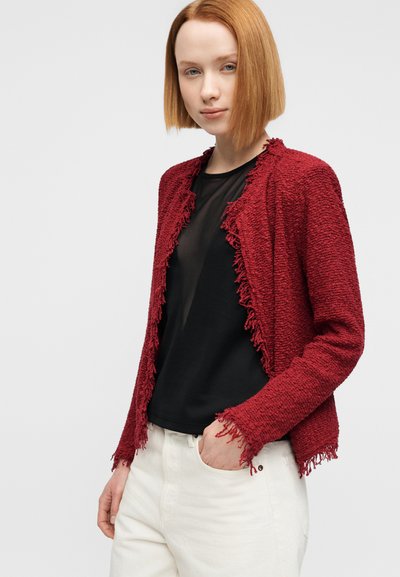 Κόκκινο πλεκτό καρό με υφή και διακοσμητικά κρόσσια στις άκρες, φορεμένο πάνω από μαύρο τοπ, συνδυασμένο με παντελόνι σε κρεμ απόχρωση. Λείο, υφαντό ύφασμα.