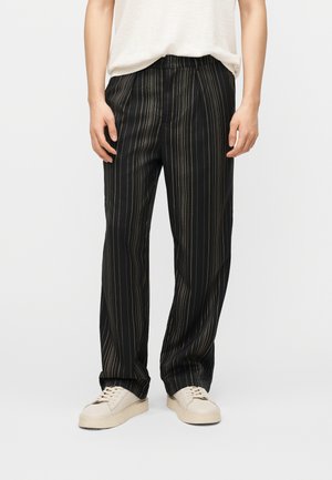 Osoba oblečená v černých kalhotách s pruhy a bílých teniskách, stojící s rukama volně podél těla, na jednoduchém světlém pozadí.