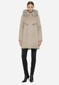 Cappotto beige con cappuccio e collo in pelliccia sintetica, vita cinchata con cordini, tasche laterali e taglio dritto, abbinato a jeans neri slim e stivali.
