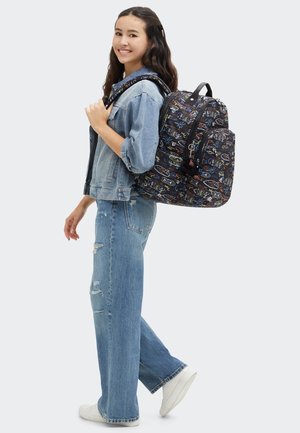 Mochila negra con gráficos coloridos, correas ajustables y un bolsillo con cremallera en la parte frontal. Llevada sobre una chaqueta de mezclilla azul y jeans desgastados.