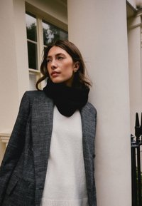 Slojeviti outfit koji uključuje teksturirani sivi sako, bijeli džemper s rebrastim rubom i crni, debeli šal. Zlatne naušnice u obliku obruča dodaju detalj.