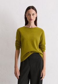 Pull en tricot vert à manches longues, avec des bords côtelés aux poignets et une encolure arrondie. Assorti à un pantalon noir ajusté. Texture lisse, coupe décontractée.