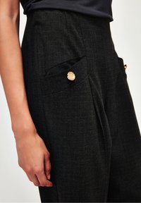 Mustat r äätälöidyt housut, joissa on värjätty kangas, laskokset ja kaksi etutaskua, joissa on kultaiset pyöreät napit.