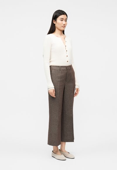 Femme aux longs cheveux noirs portant un pull crème boutonné, un pantalon large marron et des chaussures plates beiges, debout devant un fond uni.