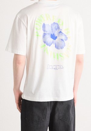 Person wearing white T-shirt with large blue flower and yellow text "POWERED BY SUNSHIN" on the back, paired with black pants.
