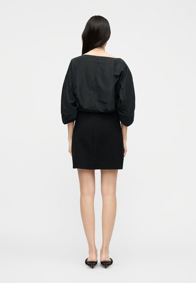 Femme aux longs cheveux noirs, portant une robe noire à manches bouffantes et des escarpins noirs à petit talon, debout de dos sur un fond uni.