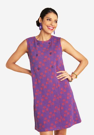 Frau lächelt, trägt ein ärmelloses lila Kleid mit rotem Blumenmuster, passende lila Ohrringe und bunte Perlenarmbänder, posiert mit den Händen in die Hüften gestützt.