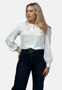 Biała bluzka z długim rękawem, z plisowanym przodem i szczegółem w kształcie klucza. Połączona z ciemnymi dżinsami i czarnym paskiem z okrągłą, złotą klamrą.