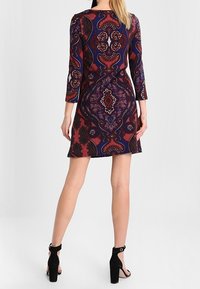 Krátký rukáv, přiléhavé šaty v odvážném paisley vzoru s hlubokými červenými, modrými a černými tóny, s kulatým výstřihem a rozšířeným spodním okrajem.