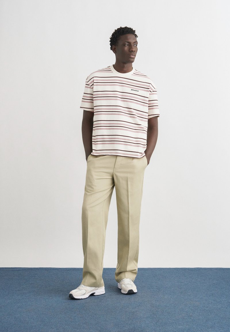 Hombre de pie con las manos en los bolsillos, vistiendo pantalones beige, camiseta blanca con rayas burdeos y zapatillas blancas sobre una alfombra azul.