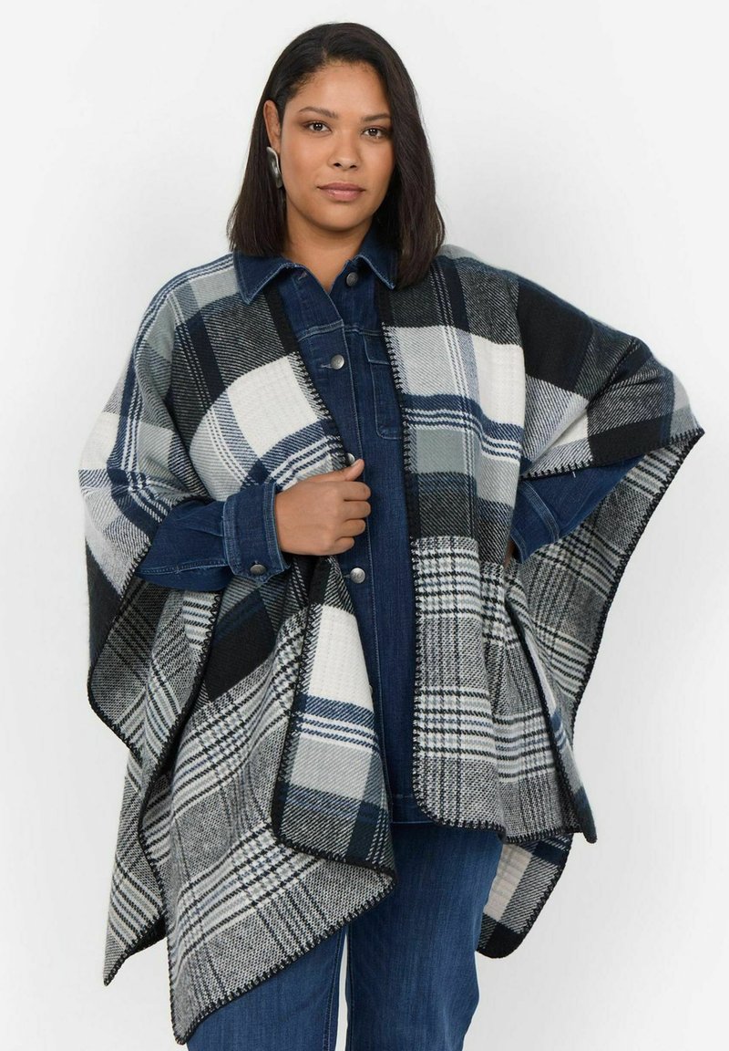 Rutig poncho i marinblått, svart och vitt. Tillverkad av mjukt, strukturerat material med fransade kanter. Bärs över en jeansskjorta.