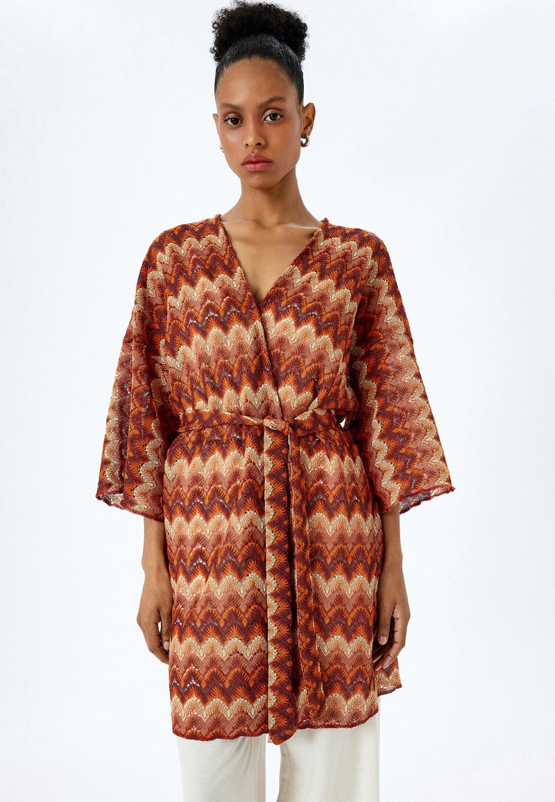 Röd och orange mönstrad kimono-stil omslag med bälte, vida ärmar och ett zick-zack mönster. Genomskinlig textur förstärker den visuella detaljeringen.