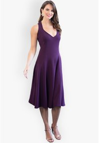 Robe sans manches violette avec un décolleté en V, jupe évasée et tissu lisse ; portée avec des talons straps en argent et des boucles d'oreilles décoratives.