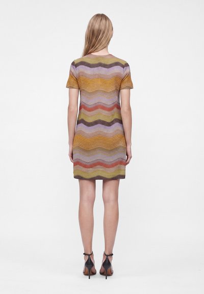 Femme aux longs cheveux blonds portant une robe courte à manches courtes, longueur genoux, avec des rayures ondulées horizontales multicolores, vue de dos.