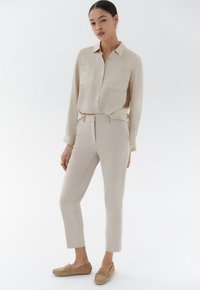 Chemise beige clair à manches longues avec deux poches poitrine, associée à un pantalon beige cintré et des chaussures mocassins marron clair.