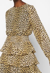 Pusero, jossa on leopardikuosi, pitkät hihat, smockattu vyötärö ja kerrokselliset röyhelöt. Kankaan kuosisävyissä hallitsevat keltaisen ja ruskean sävyt.