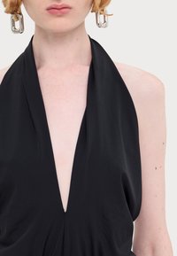 Svart halterneck-plagg med dyp V-hals, laget av myk, glatt stoff og med fremtredende draperinger. Metalliske, rektangulære øredobber fremhever stilen.