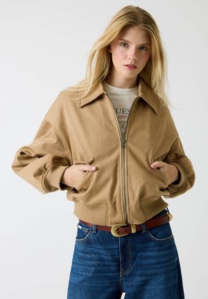 Tan bomber jacket with a front zipper, pointed collar, and ribbed cuffs. Paired with blue denim jeans and a brown belt with a circular buckle.