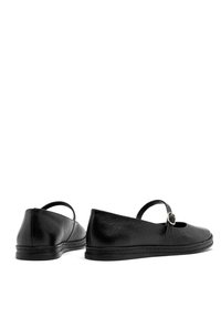 Zapatos de cuero negro con punta redondeada, suela plana y una correa decorativa con detalle de hebilla. La textura es ligeramente granulada.