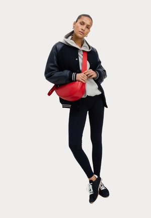Veste varsity noire et marine avec des rayures blanches, sweat à capuche gris, leggings noirs et baskets noires. Sac bandoulière rouge de forme ronde.