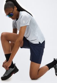 Αθλητικό άτομο γονατίζει για να δέσει γκρι και μαύρα παπούτσια πεζοπορίας, φοράει μπλε σορτς, ανοιχτό γκρι μπλούζα, μαύρες κάλτσες και αντανακλαστικά γυαλιά ηλίου.