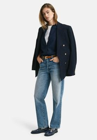 Marineblazer met gouden knopen, lichtblauwe denim jeans, bruine leren riem en marine loafers. Witte t-shirt is subtiel zichtbaar eronder.
