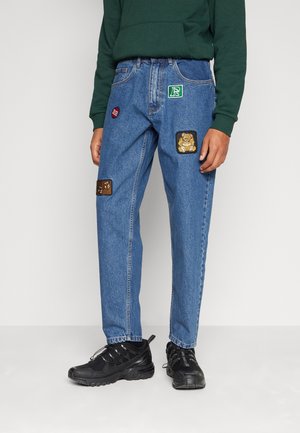 Person wearing blue denim jeans with colorful patches, black sneakers, and a dark green sweatshirt, standing against a white background.