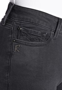 Primer plano de unos vaqueros de denim negro con un pequeño bolsillo para monedas que presenta costuras decorativas y un adorno en forma de "R" con strass cerca de la cintura.