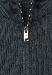 Dunkelgrauer Reißverschluss-Pullover mit gerippter Textur, metallischem Reißverschluss und hohem Kragen. Markenetikett am Reißverschluss sichtbar.