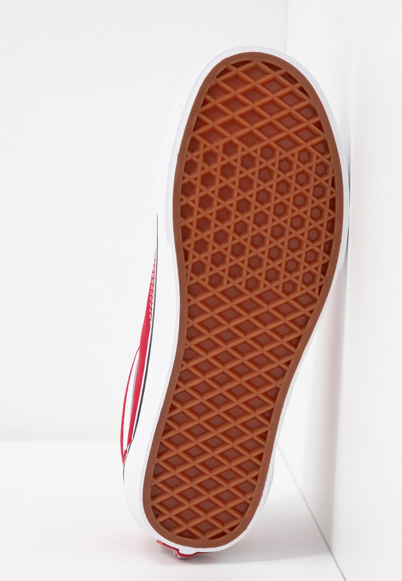 Zapatilla roja con suela de goma texturizada con un patrón de diamante, entresuela blanca y franja negra en el costado.