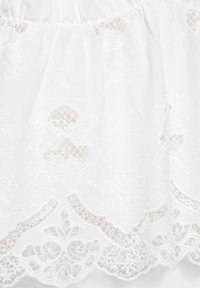 Tissu blanc avec broderie en dentelle complexe et bords festonnés, présentant des motifs découpés floraux et géométriques.