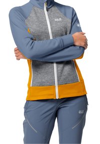 Šedá a modrá bunda s žlutými akcenty, měkký materiál, plný zip a logo na prsou. Spárována se šedými kalhotami s jemným vzorem.