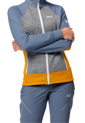 Graue und blaue Jacke mit gelben Akzenten, aus weichem Material, mit durchgehendem Reißverschluss und Logo auf der Brust. Kombiniert mit grauen Hosen mit einem dezenten Muster.