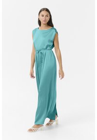 Robe sans manches turquoise avec une coupe décontractée, ceinture à la taille cintrée et fentes sur les côtés. Portée avec des sandales blanches à lanières sur un fond uni.
