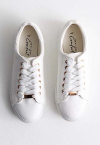 Vita canvas sneakers med platta vita snören, ögonsnören i guldton och en dekorativ guldaccent på tungan. Texturerad yta.