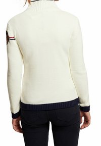 White knit sweater with a high ribbed collar, navy ribbed cuffs and hem, featuring a patch on the left arm with red and navy accents.