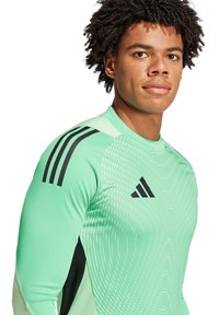 Maglietta sportiva verde a maniche lunghe con accenti neri, caratterizzata da un motivo testurizzato e tre strisce nere sulle spalle.