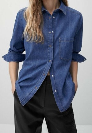 Femme portant une chemise en denim bleu boutonnée avec manches retroussées et pantalon noir, les mains dans les poches, debout devant un fond uni.