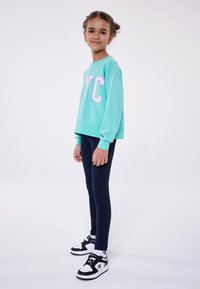 Un felpa turchese con la scritta "NYC" in lettere bianche e rosa, abbinata a leggings blu navy e sneakers in bianco e nero.