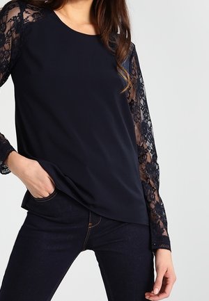 Frau trägt ein schwarzes Langarmshirt mit Spitzensäumen und dunkle Jeans, steht mit einer Hand in der Tasche vor einem einfarbigen Hintergrund.