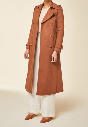 Femme portant un long trench-coat marron croisé, un pantalon blanc à jambes larges et des chaussures marron assorties à bouts pointus sur un fond neutre.