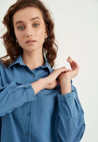 Camicia azzurra chiaro con bottoni, realizzata in un tessuto morbido, dotata di colletto, maniche lunghe e bottoni ai polsini. Il modello posa con le mani posizionate in modo elegante.