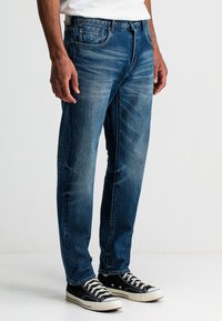 Blaue Jeans mit einem geraden Beinanschnitt, verwaschenem Look, Fünf-Taschen-Design und einem Logodetail auf der linken Vorderseite Tasche. Kombiniert mit schwarzen Sneakern.
